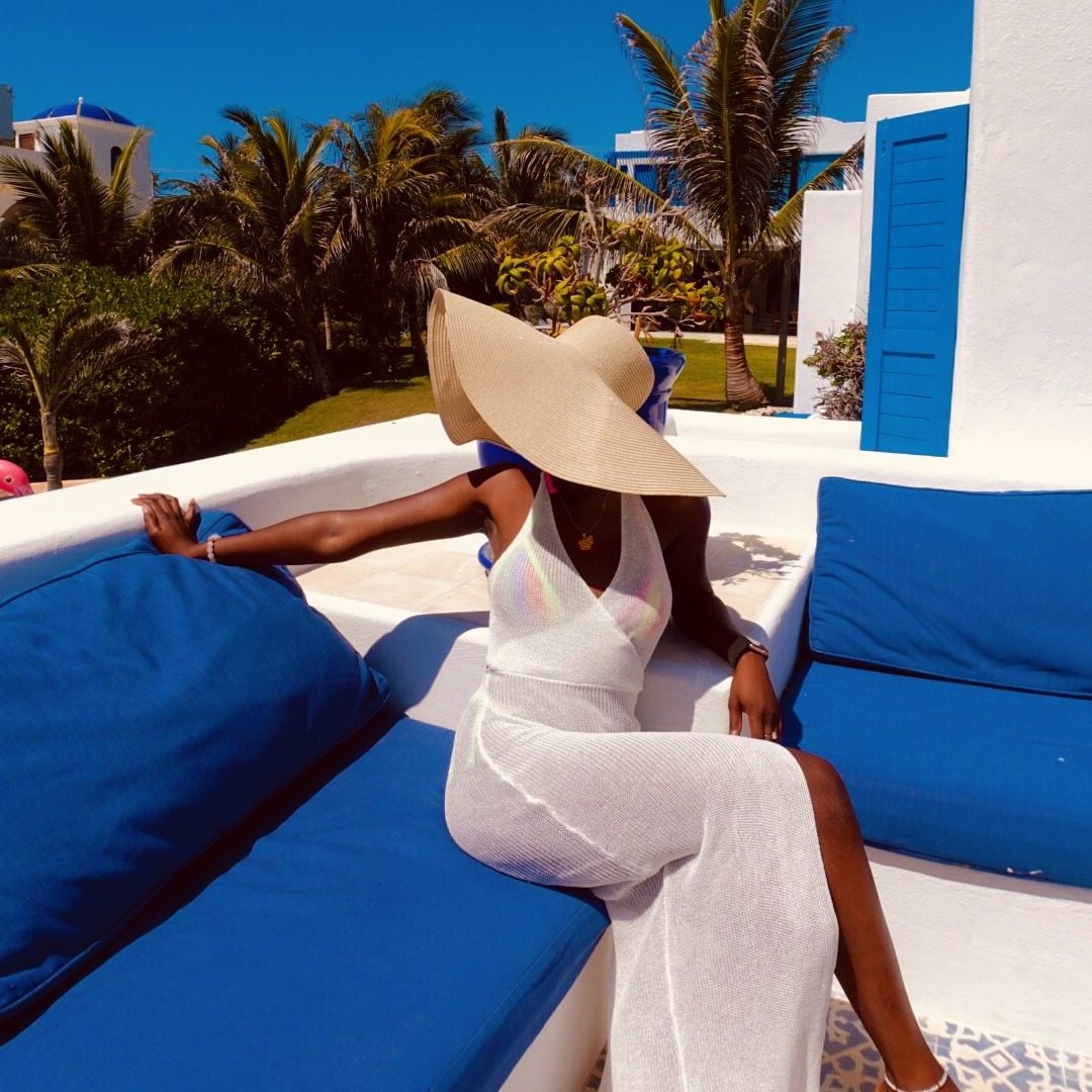Black girl in a white knitted dress with the sun hat sitting on a blue chair with blue skies in Mexico