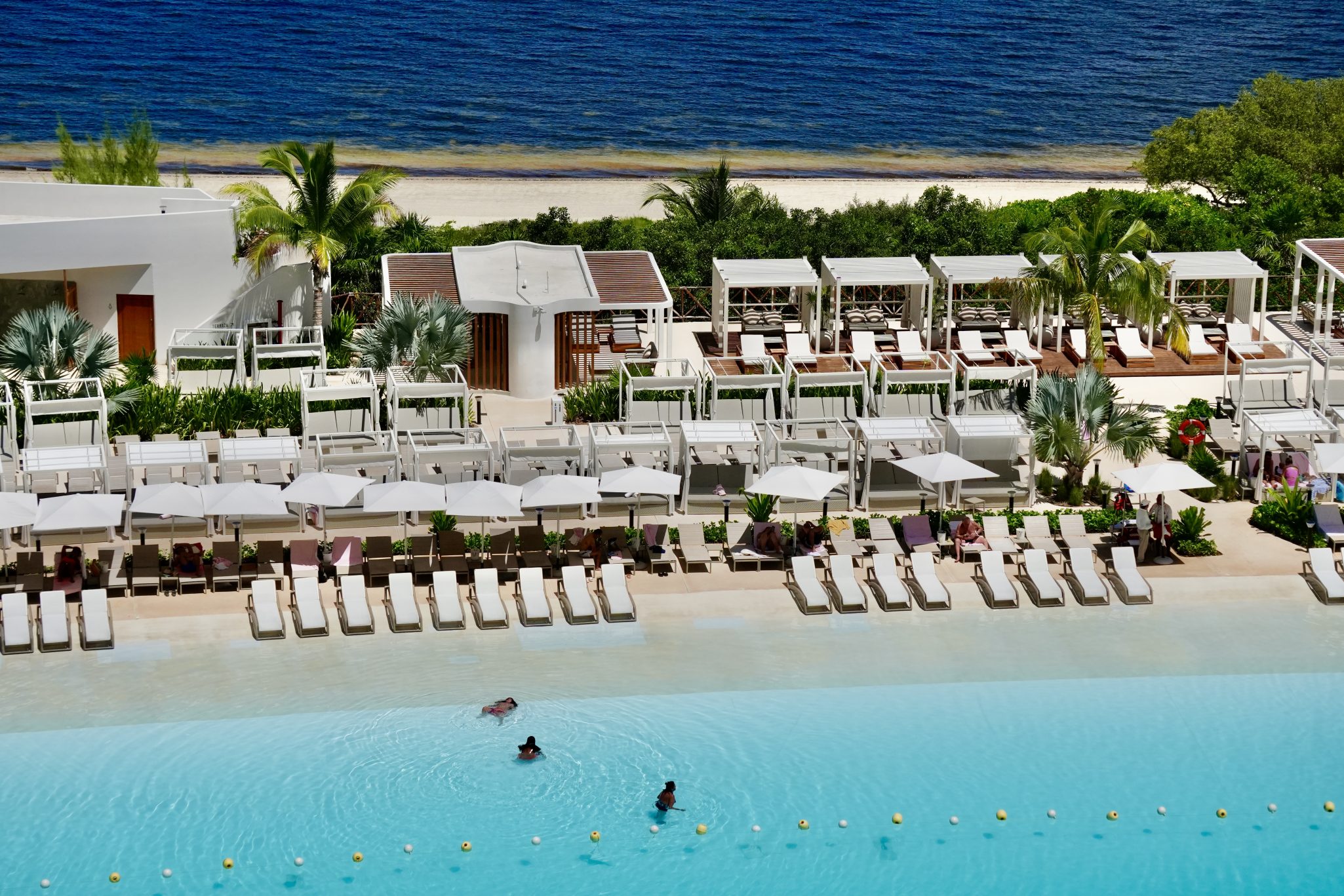 A luxury beachfront resort with rows of white lounge chairs and umbrellas surrounding a large swimming pool. Private cabanas with white canopies line the background, with lush palm trees and greenery leading to a sandy beach and the blue ocean beyond. Guests are swimming in the pool and relaxing in shaded seating areas.