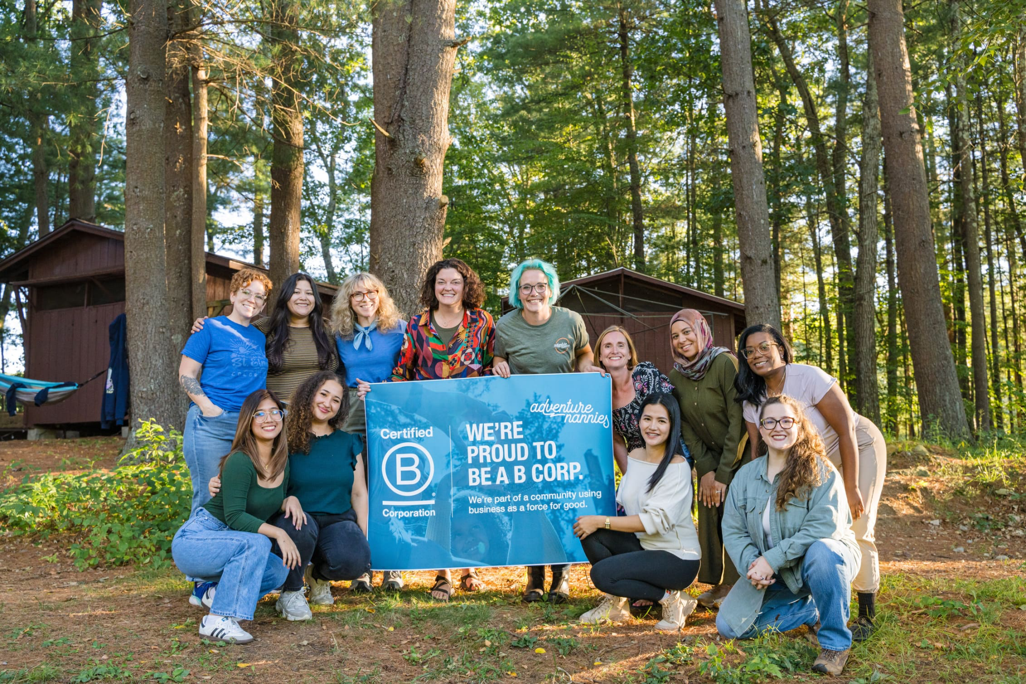Group of individuals posing together outdoors, holding a sign that reads 'WE'RE PROUD TO BE A B CORP.' surrounded by trees and cabins.