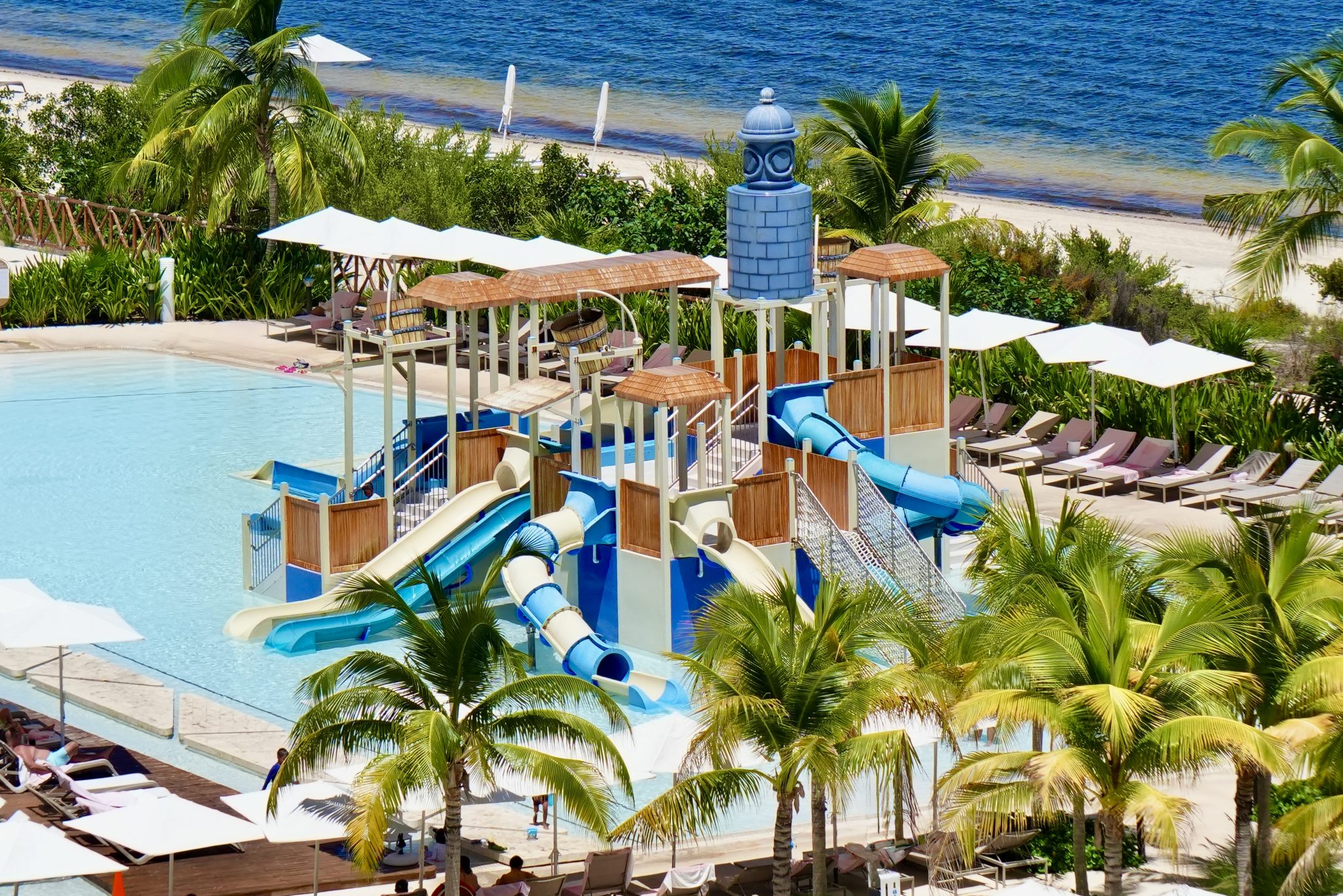 A colorful water slide attraction at a resort pool, surrounded by palm trees and lounge chairs, with a beach and ocean in the background.