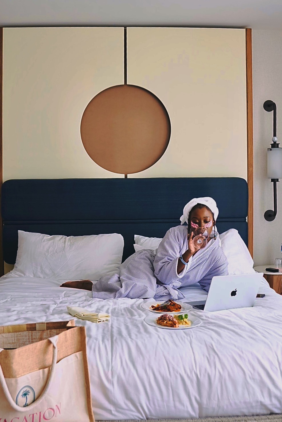 A woman in a striped bathrobe lounging on a hotel bed, drinking from a glass while working on a laptop, with a plate of food beside her.