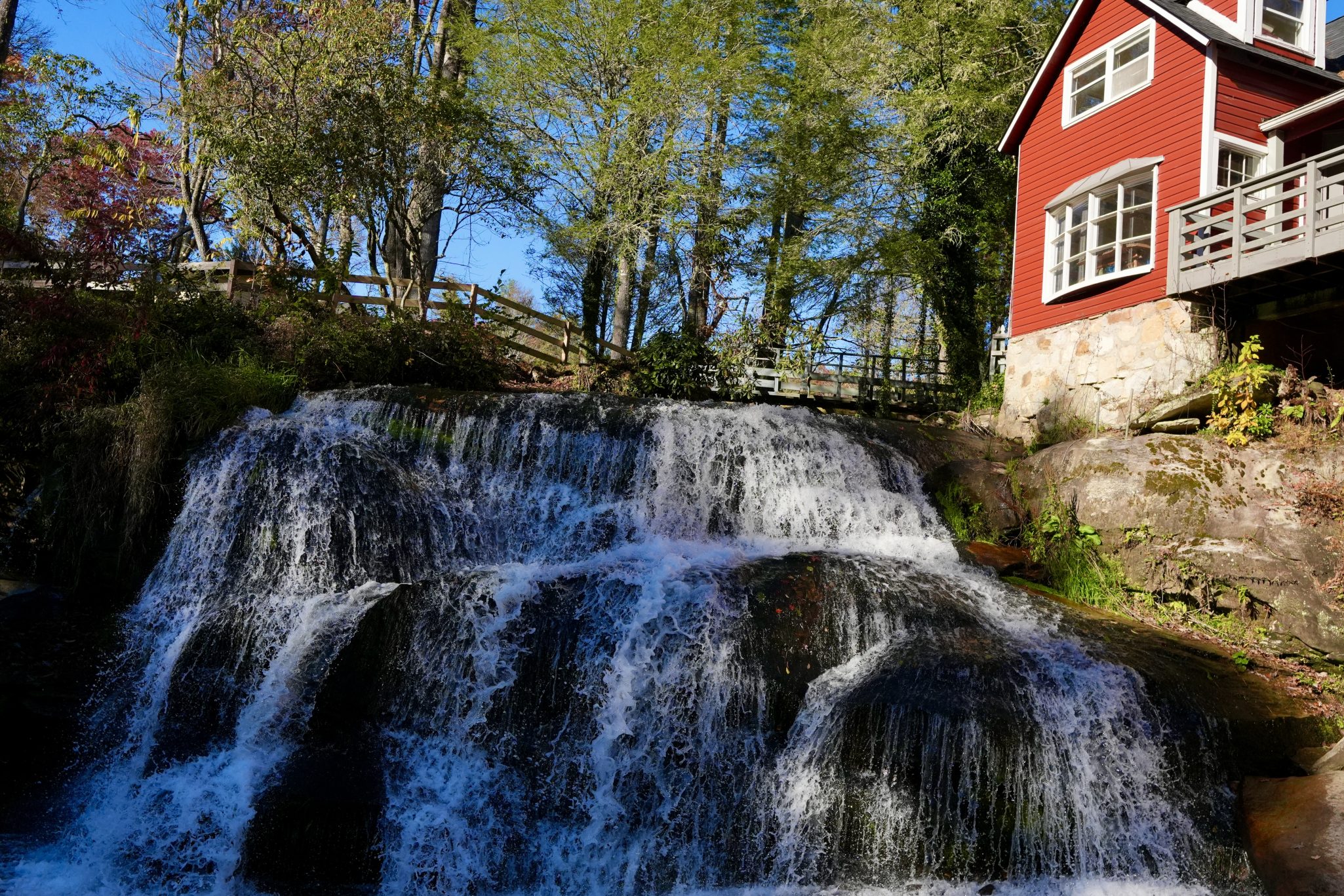 A picturesque waterfall cascading over rocks, with a red cabin visible in the background, surrounded by trees and a clear blue sky.