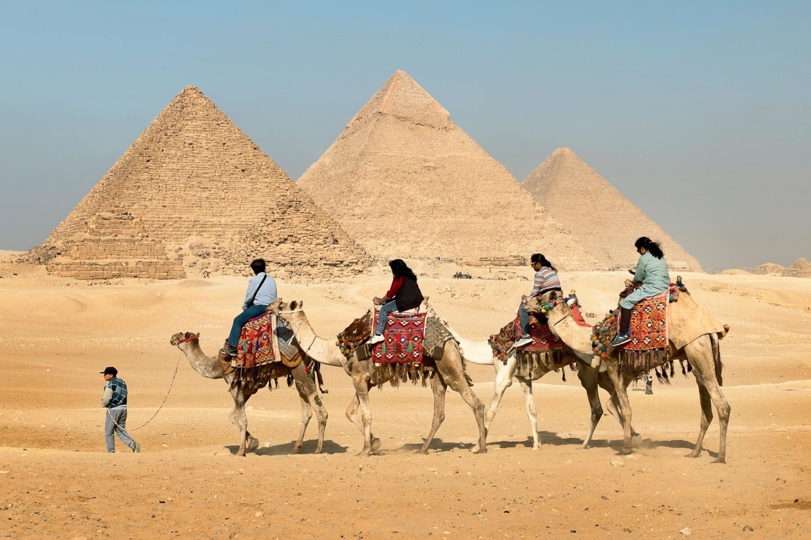 Four people riding camels in the sandy desert near the Pyramids of Giza, with a guide walking alongside.