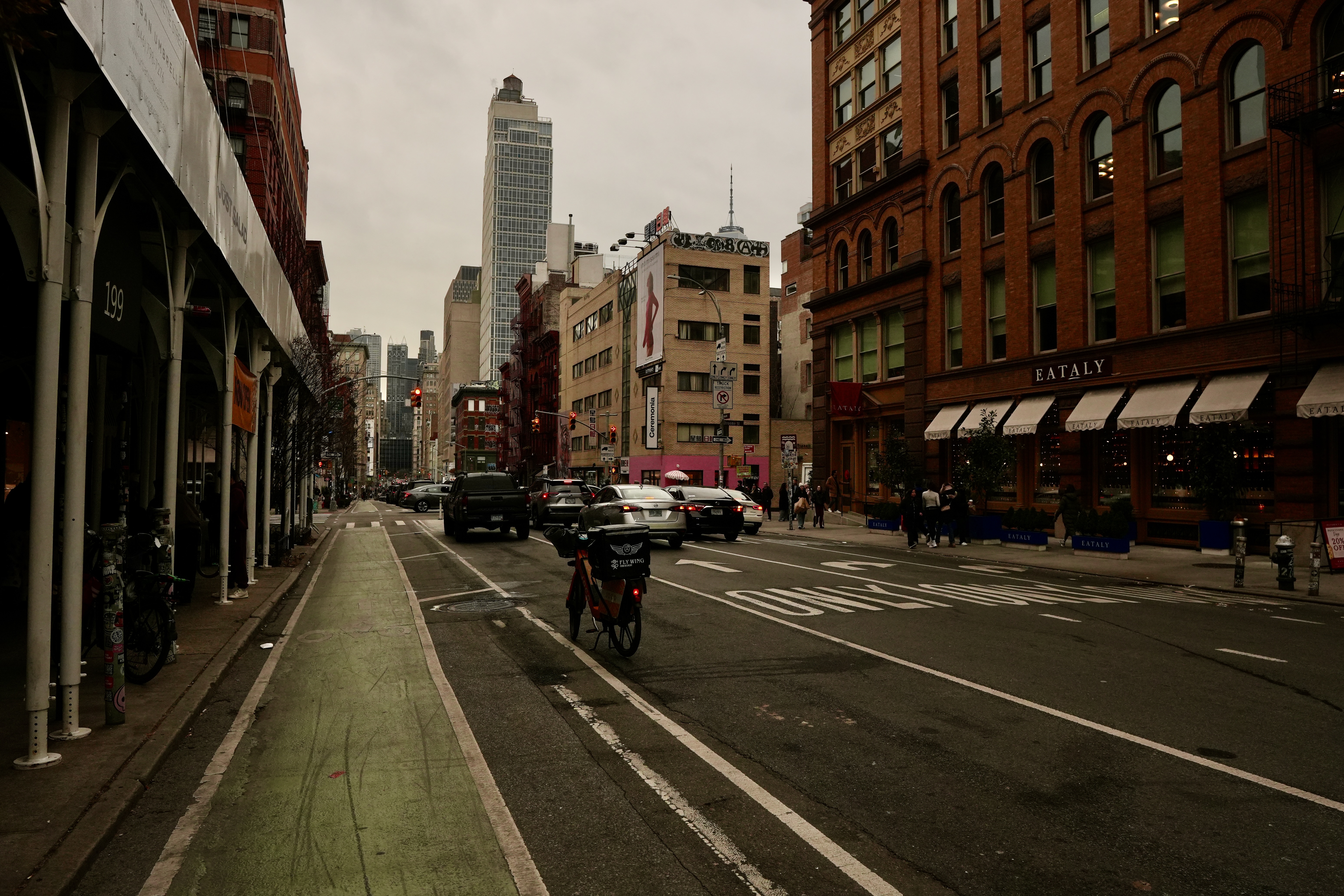 A busy urban street scene with a bike lane, featuring vehicles and pedestrians amidst buildings, including a restaurant with outdoor seating.