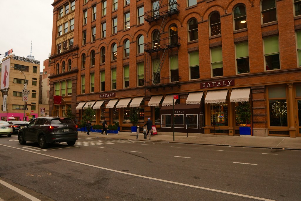 A street view of Eataly, a bustling Italian marketplace, featuring a brick building façade with large windows. Cars and pedestrians are present on the urban street.