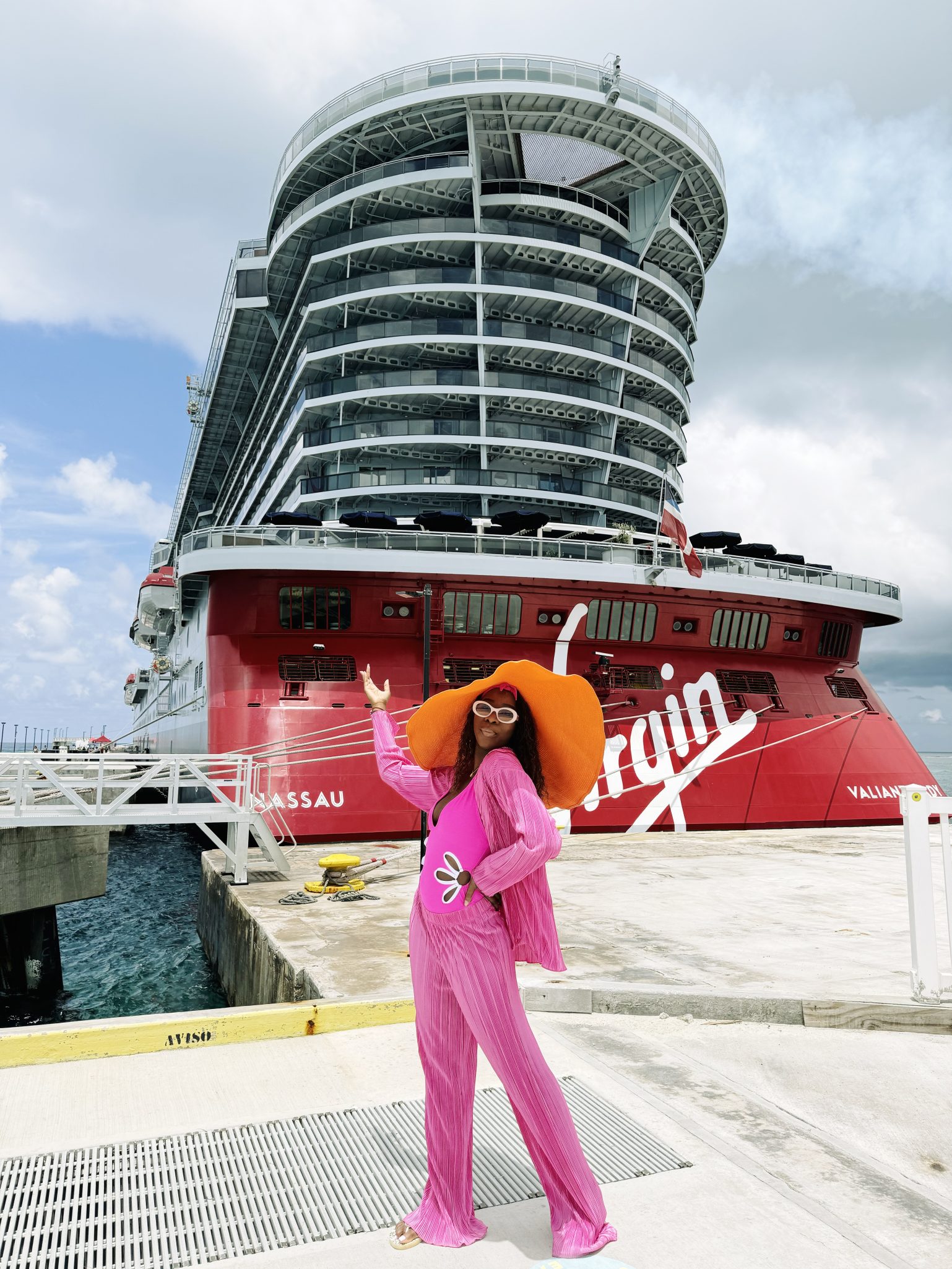 A person wearing a bright pink outfit and a large orange hat poses in front of a Virgin Voyages cruise ship docked at a port.