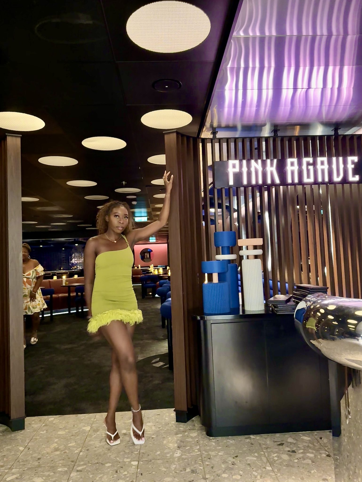 A stylish person posing in a vibrant yellow dress with a feathered hemline, standing in a modern restaurant setting featuring illuminated signage that reads 'PINK AGAVE'.