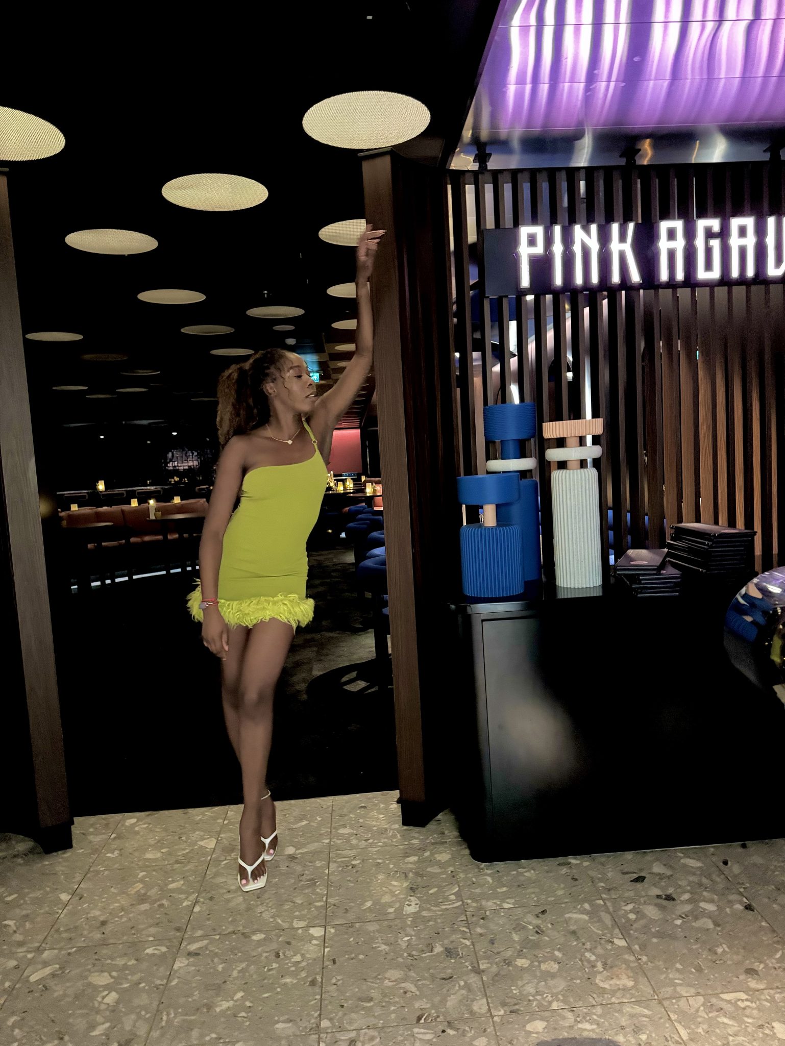 A person in a bright yellow dress with a feathered hem stands in a stylish interior space with modern decor, posing near a sign that reads 'PINK AGAVE'.