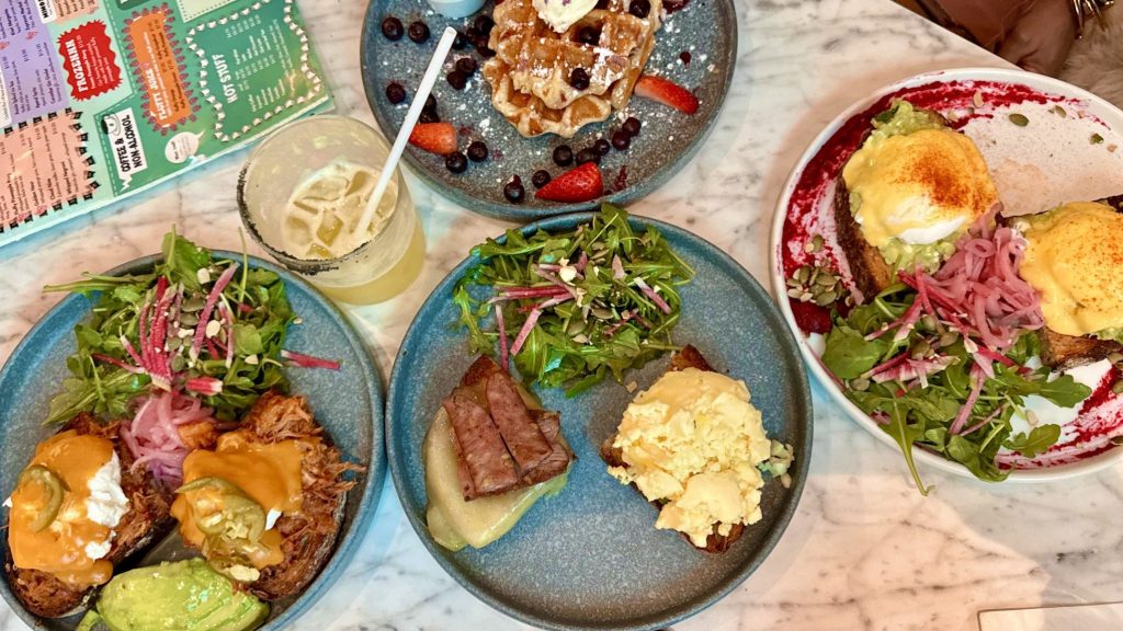 A spread of brunch dishes on a marble table, including avocado toast, eggs, salad, and waffles topped with fruit.
