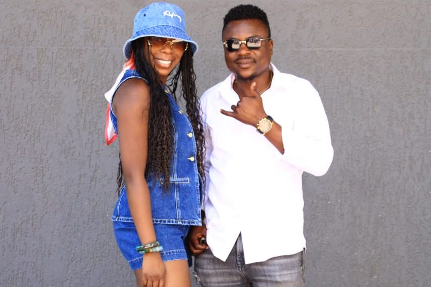 A smiling woman with long braids wearing a denim outfit and blue bucket hat stands next to a man in a white shirt and sunglasses, both posing playfully against a gray background.
