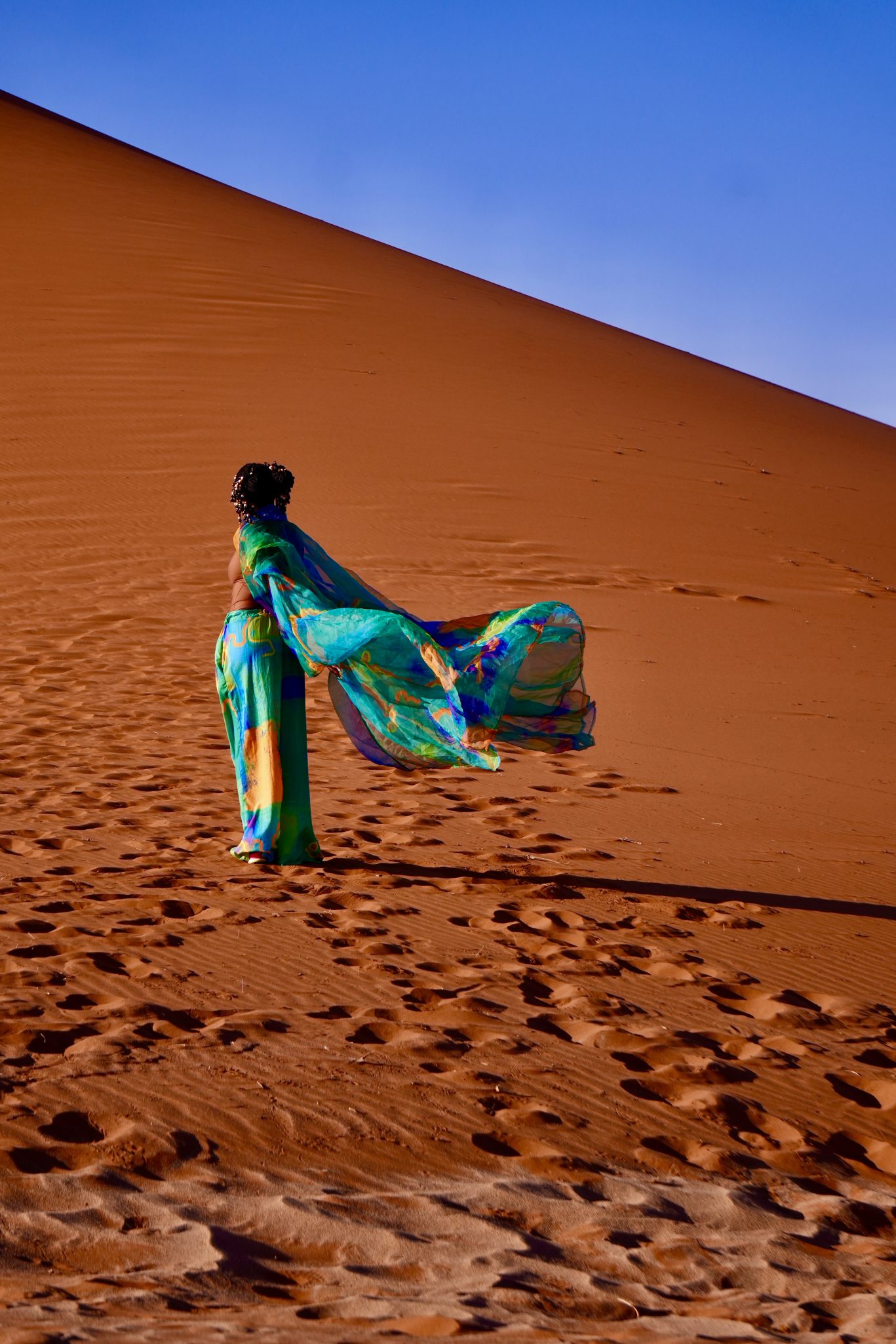 A person wearing a colorful outfit stands in a desert, with a large sand dune in the background and a blue sky above. The outfit includes a flowing cape that billows in the wind.