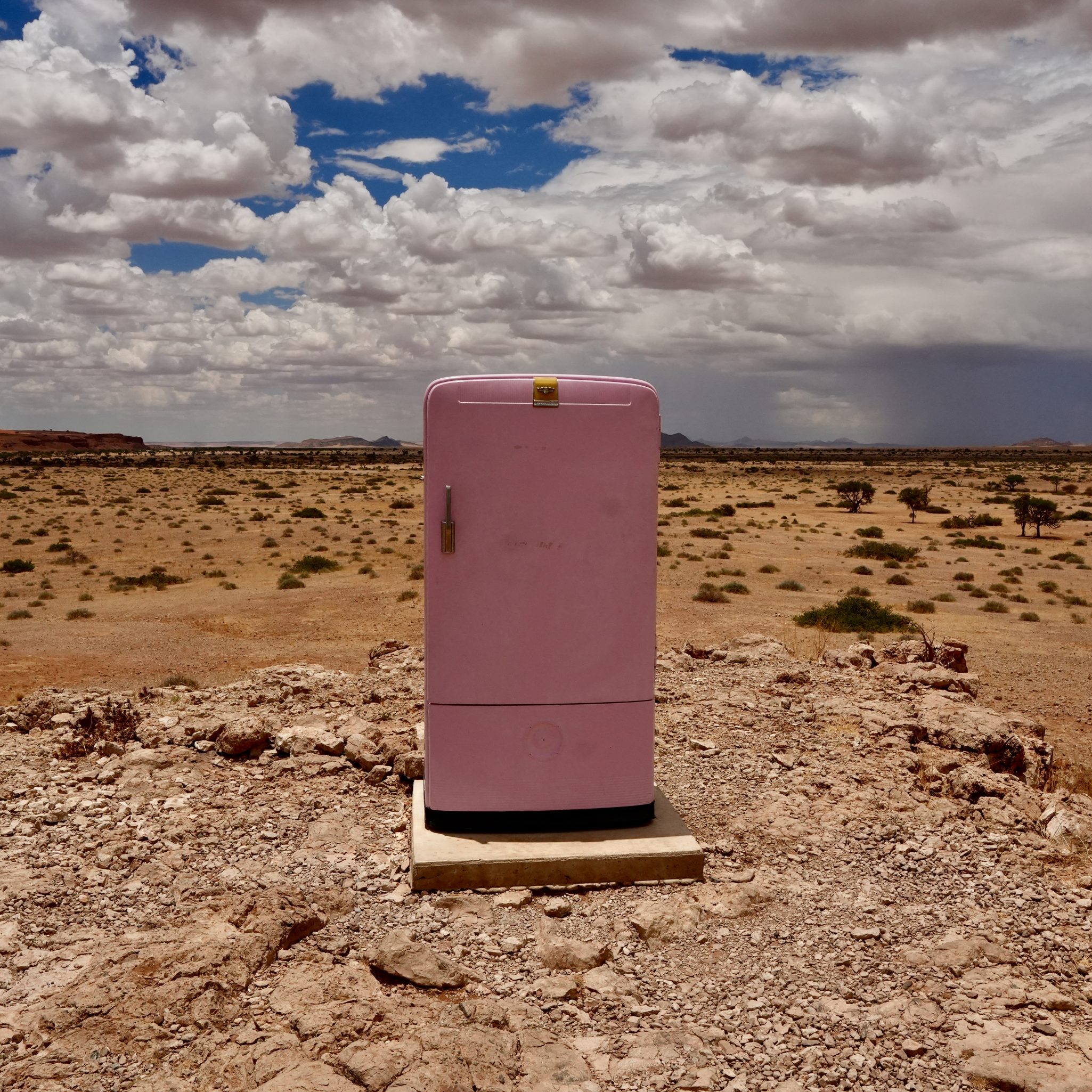 A pink refrigerator standing alone on a rocky surface in a vast desert landscape under a cloudy sky.