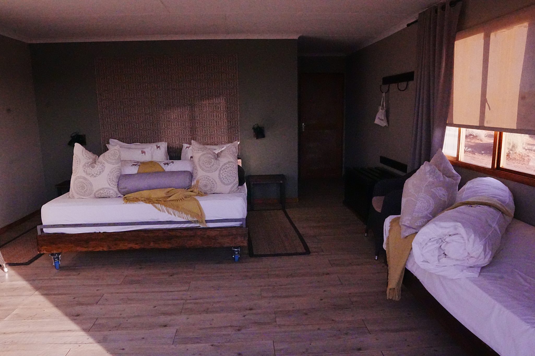 Interior of a cozy room featuring a double bed with decorative pillows, a gray blanket, and a yellow throw. There's a side table, a single bed with rolled linens, and a window with natural light filtering in.