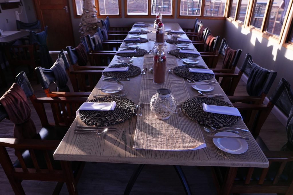 A long dining table set for a meal, featuring woven placemats, white napkins, and cutlery, with decorative bottles and a soft ambiance.