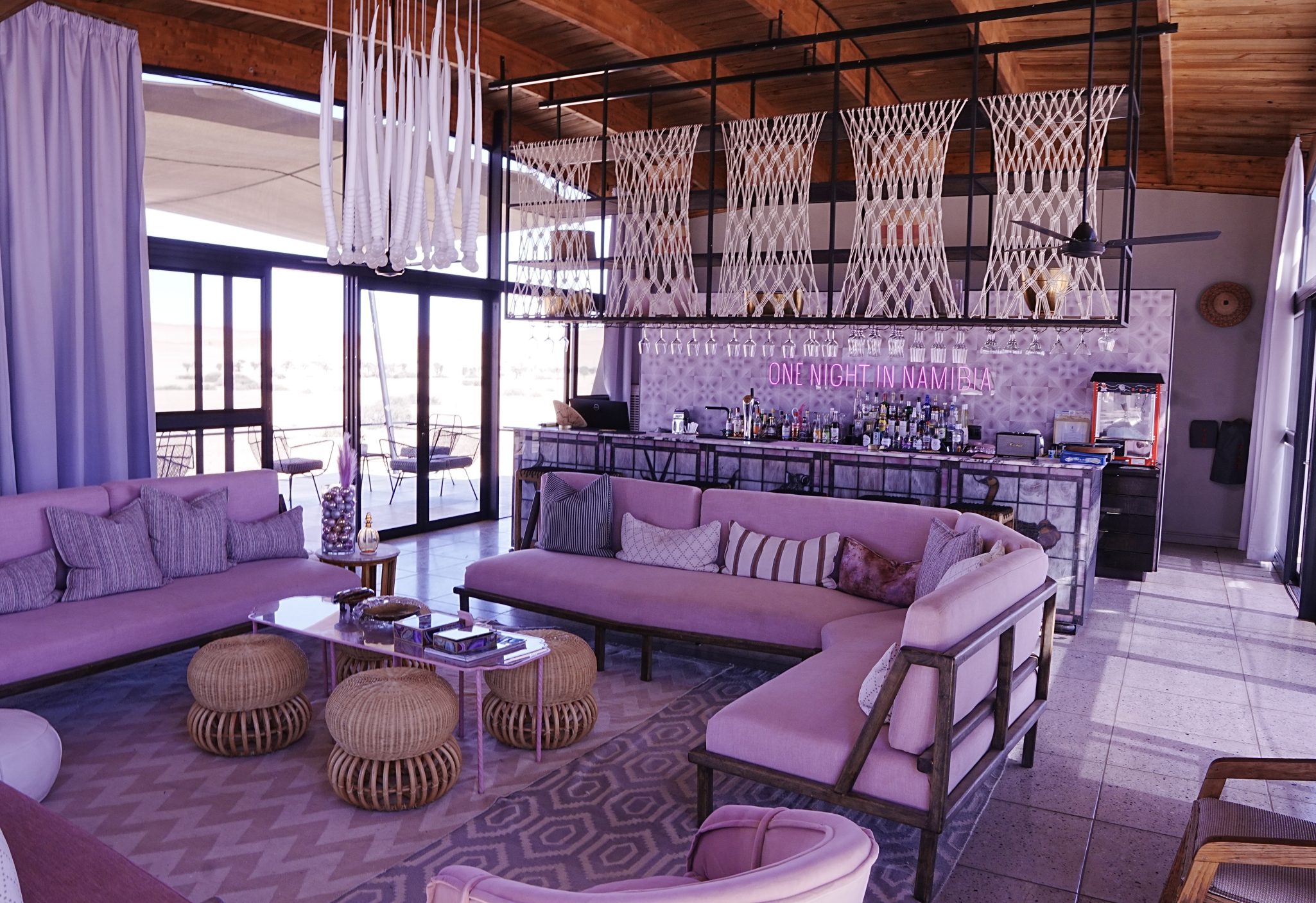A stylish indoor lounge area featuring pink sofas, round wicker tables, and decorative cushions. The backdrop includes a bar with a variety of bottles and a large window offering a view of the outdoor setting.