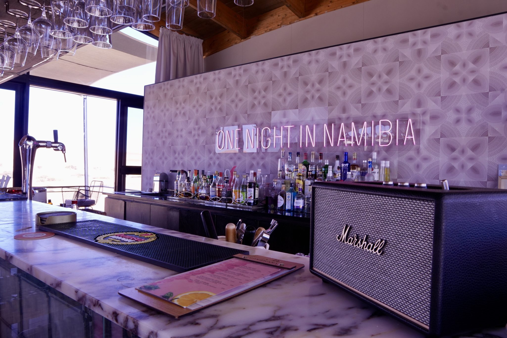 A stylish bar area featuring a marble countertop, a variety of liquor bottles on shelves, and a decorative neon sign that reads 'ONE NIGHT IN NAMIBIA'. A Marshall speaker is placed on the right.