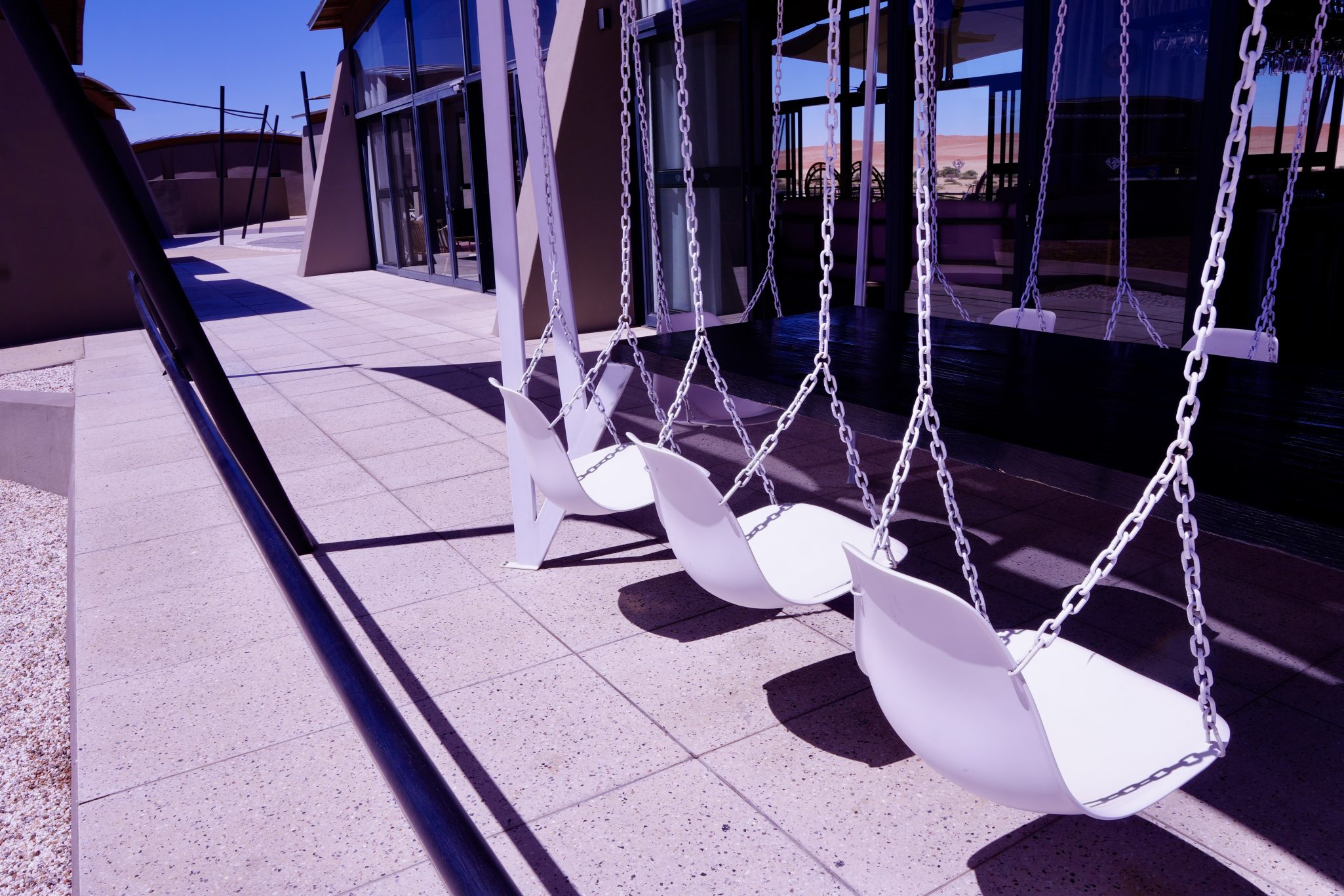 A row of modern white swings hanging from a metal frame, set against a backdrop of a contemporary building and a clear blue sky.