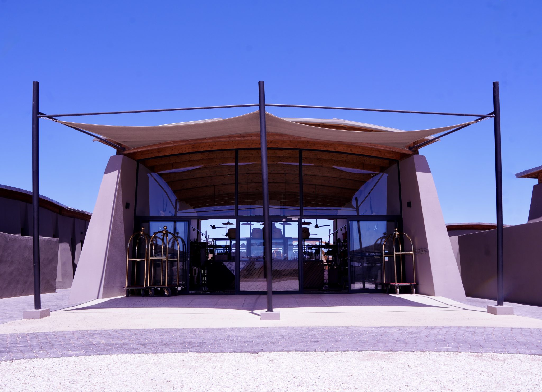 Modern architectural entrance of a building with large glass doors and a distinctive curved roof, set against a clear blue sky.