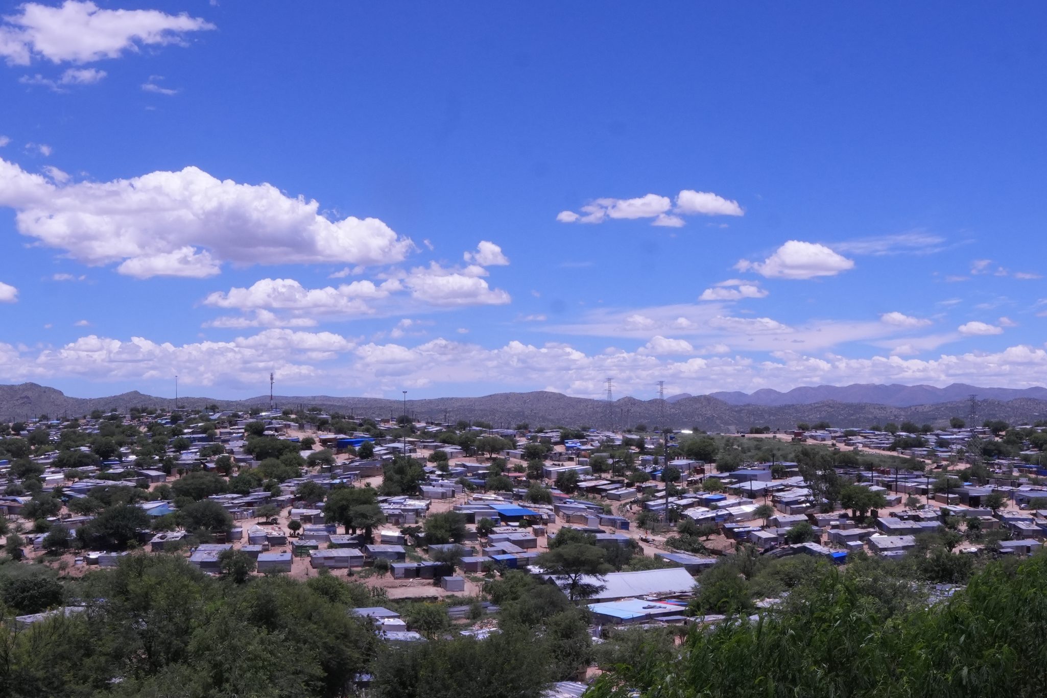 A panoramic view of a hillside community with numerous small structures under a bright blue sky adorned with fluffy clouds, surrounded by mountains.