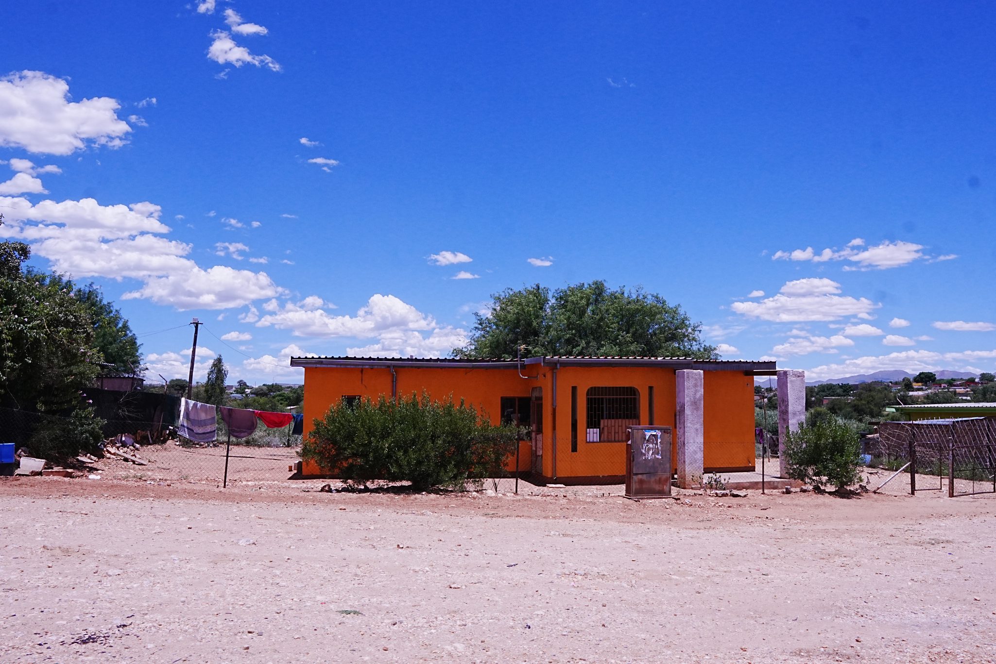 A vibrant orange house with a corrugated roof surrounded by shrubs, set against a bright blue sky dotted with fluffy white clouds. Clothes hang on a line to the left of the house, and the ground is sandy and barren.