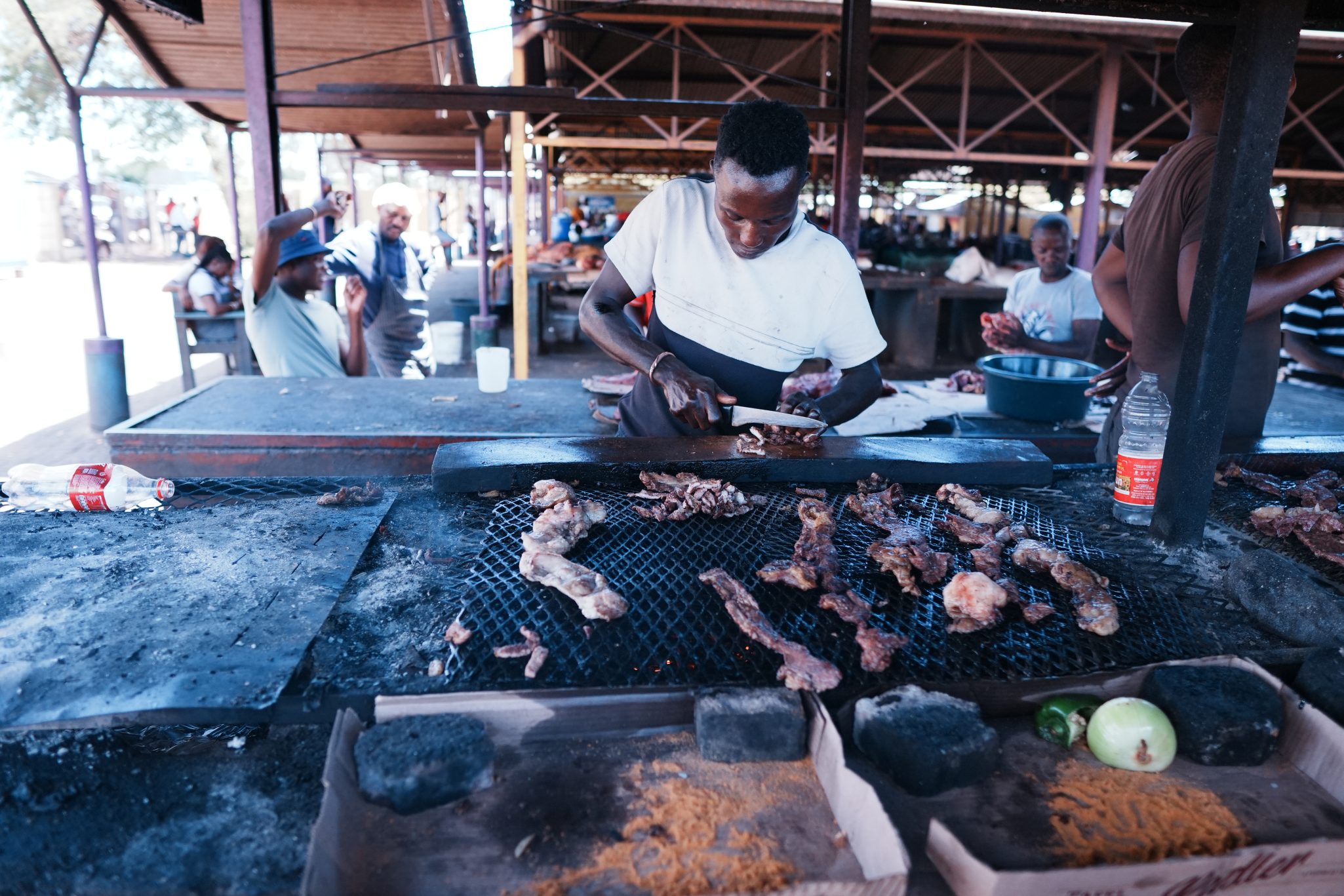 A food vendor grilling various cuts of meat over an open flame at a market. In the background, other customers and vendors can be seen socializing and preparing food.