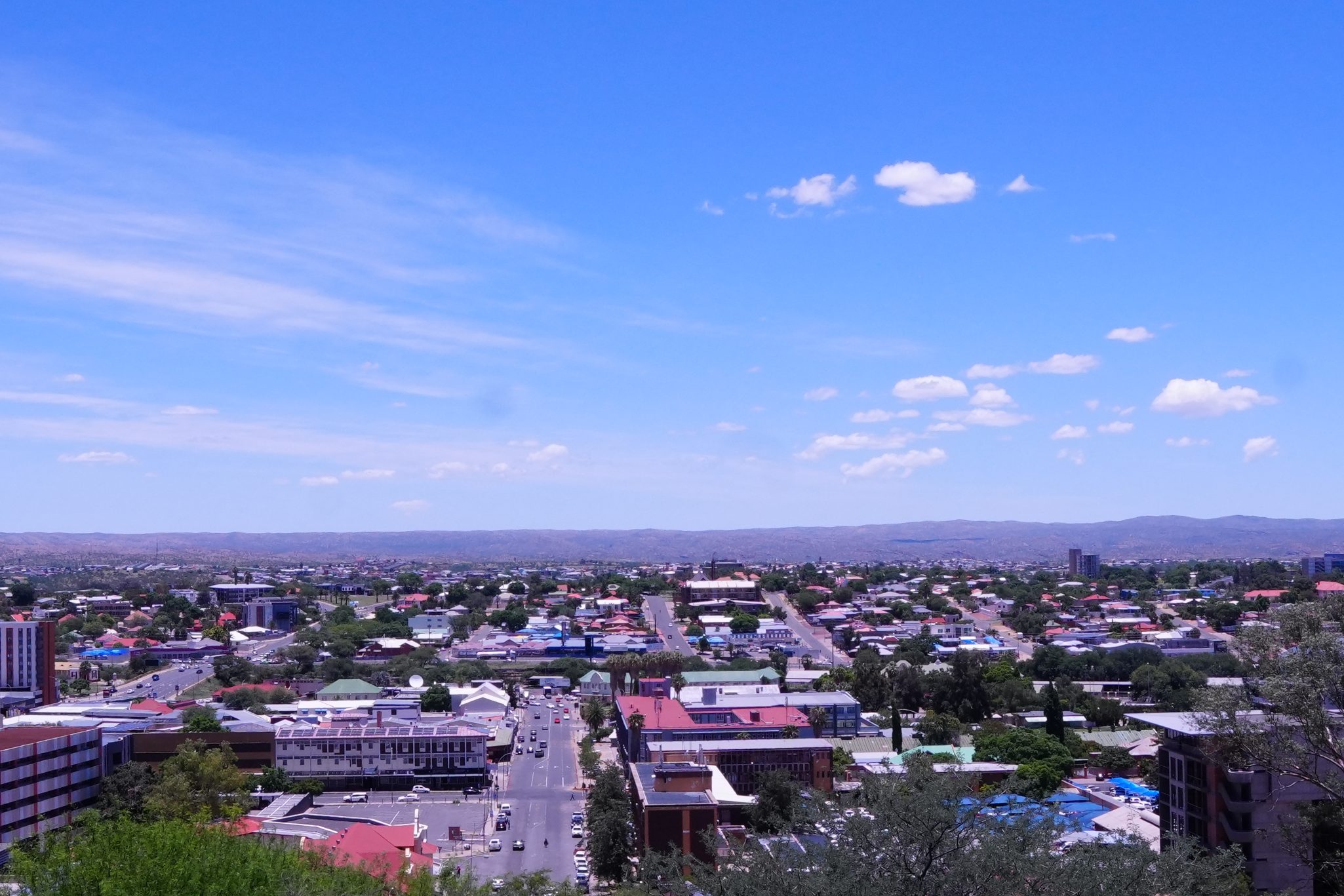 A panoramic view of a city under a bright blue sky with scattered clouds and distant mountains, showcasing various buildings and streets lined with greenery.