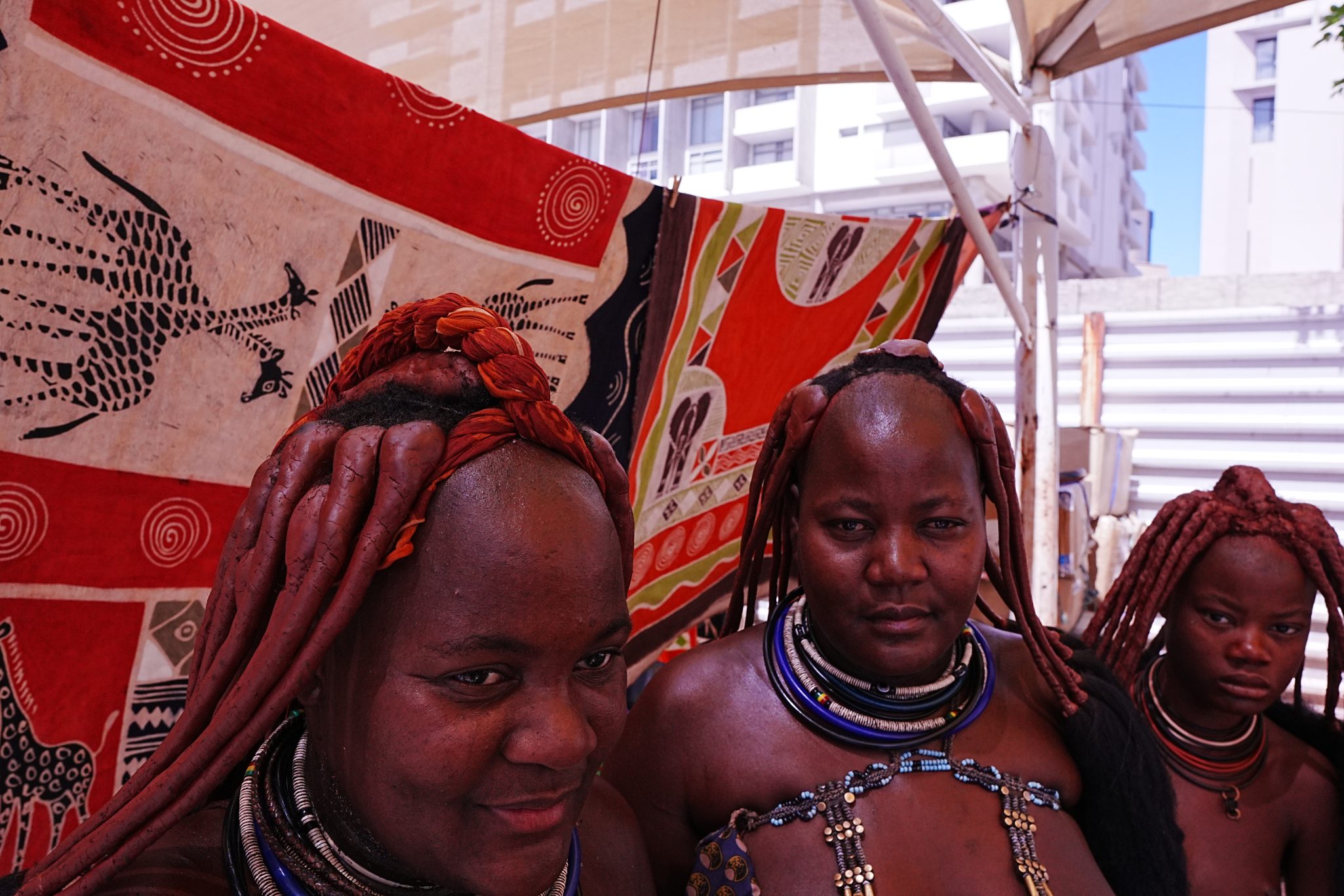 Three women dressed in traditional attire with intricate hairstyles, standing in front of patterned textiles.