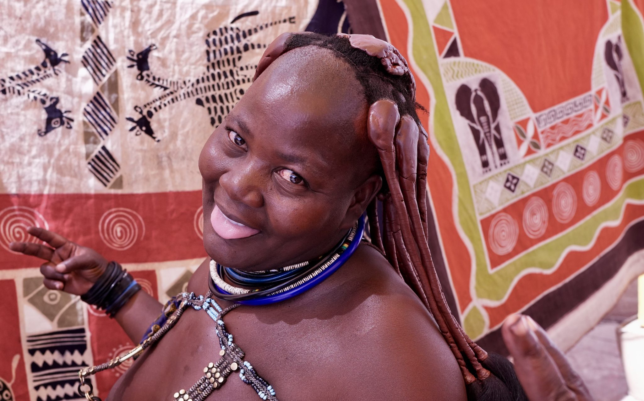 A smiling person with braided hair poses playfully while wearing colorful jewelry against a backdrop of patterned fabric.