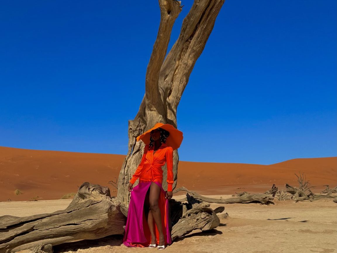 A woman in an orange outfit and large hat stands next to a gnarled tree in a desert setting with a blue sky and orange sand dunes in the background.