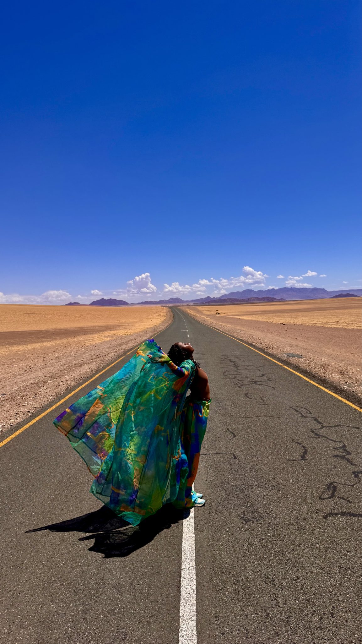 A person wearing a colorful flowing garment stands on a deserted road, arms outstretched, with a blue sky and distant mountains in the background.