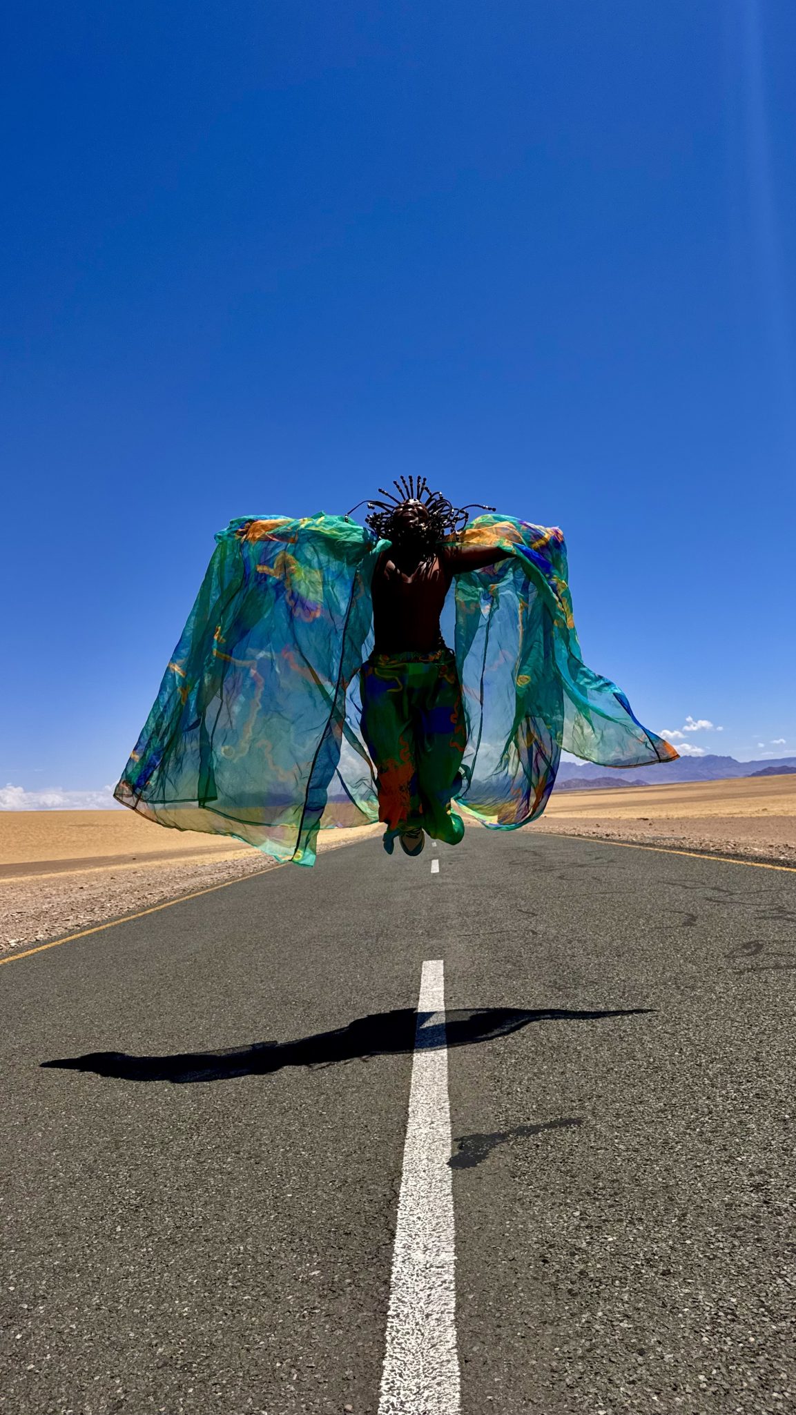 A person in vibrant, flowing clothing is leaping into the air on a deserted road under a clear blue sky, showcasing a colorful fabric backdrop.
