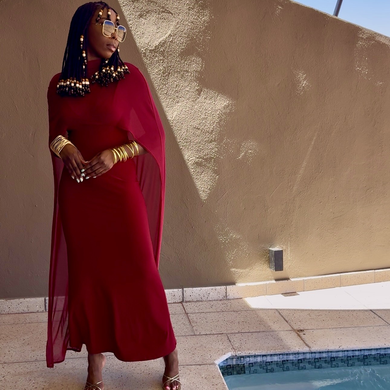 A woman poses elegantly by a pool, wearing a long red dress with a flowing cape and striking accessories, including sunglasses and bold jewelry.
