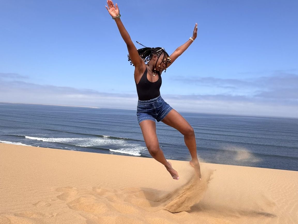Person jumping joyfully on a sandy dune with the ocean in the background.