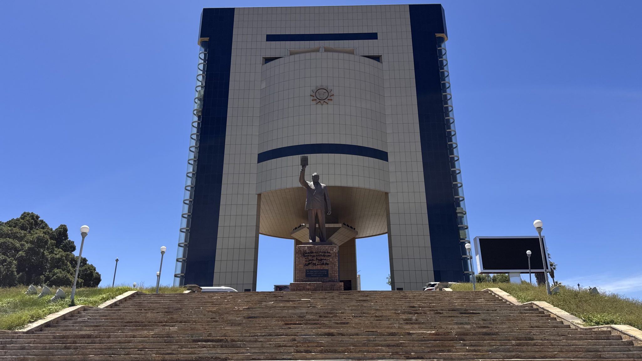 A large modern building with a prominent statue of a person holding a book at the entrance, surrounded by stairs and greenery under a clear blue sky.